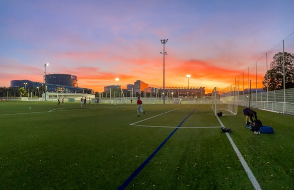 A soccer field is depicted during sunset, with an expansive sky featuring vibrant orange, pink, and purple hues. Players are scattered across the field, some practicing and others standing near the goalposts. The field is enclosed by a white fence, and in the background, modern buildings and trees are visible, illuminated by artificial lights.