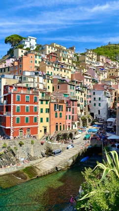 A vibrant coastal village characterized by its colorful, stacked buildings on a hillside. The scene includes a small harbor with clear turquoise water and boats moored alongside. People are leisurely walking around, enjoying the sunny day.