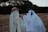 Two figures are covered head to toe in white sheets with eye holes cut out, resembling ghosts. One ghost is wearing a straw hat and sunglasses, holding a small cup. The second ghost has headphones on. They are seated in a field among cut hay or straw bales, with a line of trees in the background.