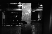 A dark and moody outdoor or subway setting with heavy shadows. The reflection of a person taking a photograph is visible in the glass, adding a sense of mystery. The lighting highlights the texture of the wall and creates an eerie atmosphere.