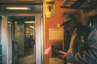 A person wearing a cap and jacket is standing inside a train compartment, looking down at a device in their hand. The train interior features metallic surfaces, signs, and lighting, contributing to a warm and slightly dim ambiance.