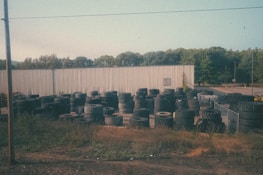 A large number of tires are stacked and scattered around an outdoor area with an industrial-looking metal building in the background. The setting includes overgrown grass and weeds around the edges, and several trees are visible beyond the building. The sky is clear with a pale blue hue.