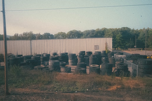 A large number of tires are stacked and scattered around an outdoor area with an industrial-looking metal building in the background. The setting includes overgrown grass and weeds around the edges, and several trees are visible beyond the building. The sky is clear with a pale blue hue.