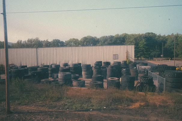 A large number of tires are stacked and scattered around an outdoor area with an industrial-looking metal building in the background. The setting includes overgrown grass and weeds around the edges, and several trees are visible beyond the building. The sky is clear with a pale blue hue.