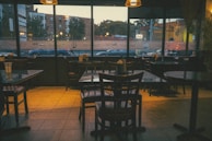 A quiet, dimly lit interior of a restaurant with several empty wooden tables and chairs. The large windows reveal an urban street outside with buildings and parked cars visible. There are condiments placed on the tables, and the ambient lighting gives the space a warm, cozy atmosphere.