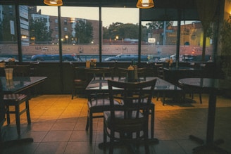 A quiet, dimly lit interior of a restaurant with several empty wooden tables and chairs. The large windows reveal an urban street outside with buildings and parked cars visible. There are condiments placed on the tables, and the ambient lighting gives the space a warm, cozy atmosphere.