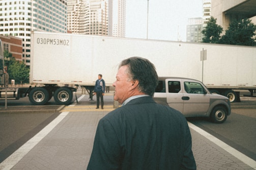 A man in a suit is seen walking on a city street corner, with a large truck and an SUV nearby. Another man stands in front of the truck, which is parked across the intersection. Skyscrapers in the background suggest an urban setting.