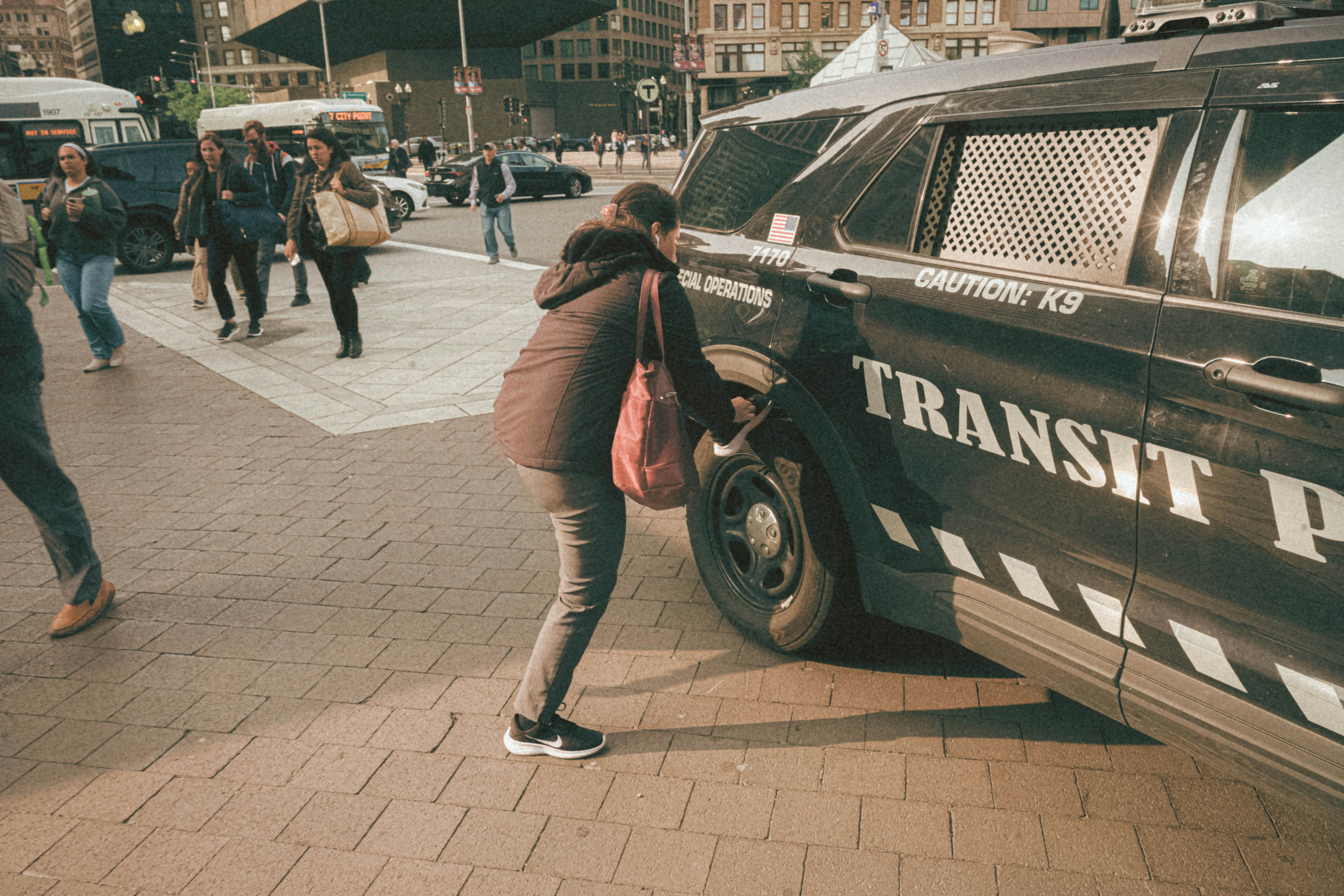 A woman is crouching beside a transit police vehicle parked on a busy city street. Other people are walking nearby, some appearing to cross the street. The environment suggests an urban setting with buildings and traffic visible in the background. The vehicle has markings indicating special operations and caution for a K9 unit.