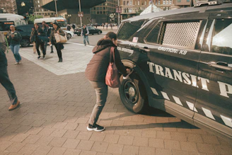 A woman is crouching beside a transit police vehicle parked on a busy city street. Other people are walking nearby, some appearing to cross the street. The environment suggests an urban setting with buildings and traffic visible in the background. The vehicle has markings indicating special operations and caution for a K9 unit.