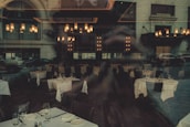 A dimly lit restaurant interior with elegantly set tables, featuring white tablecloths and neatly arranged glassware. The chandeliers provide warm lighting, enhancing the upscale ambiance. Reflections from the window showcase cars and the exterior of a building, adding an urban twist.