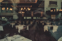 A dimly lit restaurant interior with elegantly set tables, featuring white tablecloths and neatly arranged glassware. The chandeliers provide warm lighting, enhancing the upscale ambiance. Reflections from the window showcase cars and the exterior of a building, adding an urban twist.