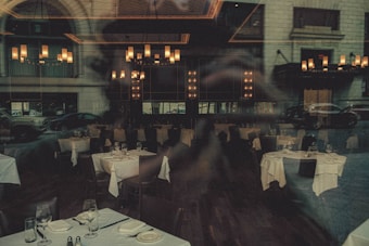 A dimly lit restaurant interior with elegantly set tables, featuring white tablecloths and neatly arranged glassware. The chandeliers provide warm lighting, enhancing the upscale ambiance. Reflections from the window showcase cars and the exterior of a building, adding an urban twist.