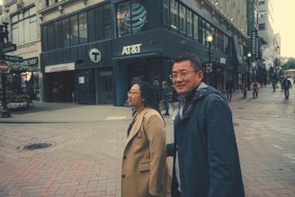 Two people are walking along a bustling city street lined with various shops and businesses, including AT&T and Starbucks. They appear to be engaged in conversation as other pedestrians move about in the background.