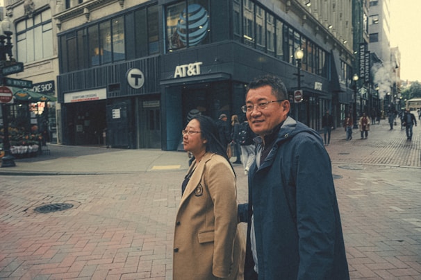 Two people are walking along a bustling city street lined with various shops and businesses, including AT&T and Starbucks. They appear to be engaged in conversation as other pedestrians move about in the background.