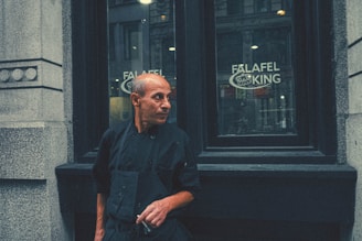 A person stands in front of a restaurant window with the sign 'Falafel King' visible. The individual is dressed in a black uniform, likely indicating their role as a chef or staff member. The urban setting and architectural details suggest a city environment. The person's expression appears thoughtful or contemplative.