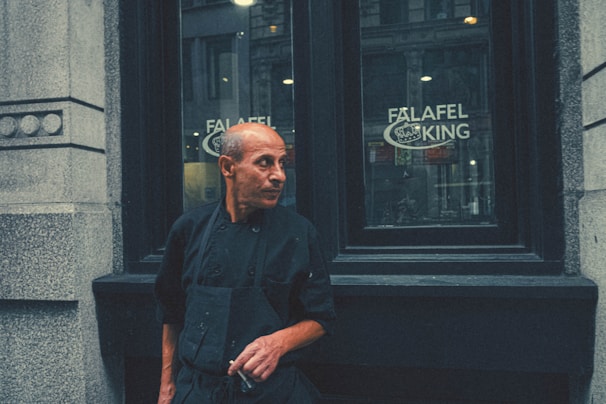 A person stands in front of a restaurant window with the sign 'Falafel King' visible. The individual is dressed in a black uniform, likely indicating their role as a chef or staff member. The urban setting and architectural details suggest a city environment. The person's expression appears thoughtful or contemplative.