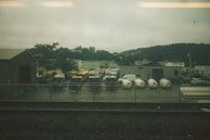 A fenced industrial area with several parked vehicles, including school buses, a FedEx truck, and white delivery vans. There are also utility trailers and equipment enclosed in tarp materials. Surrounding this are various buildings, including a warehouse, against a backdrop of green hills under an overcast sky.