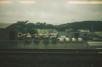 A fenced industrial area with several parked vehicles, including school buses, a FedEx truck, and white delivery vans. There are also utility trailers and equipment enclosed in tarp materials. Surrounding this are various buildings, including a warehouse, against a backdrop of green hills under an overcast sky.