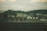 A fenced industrial area with several parked vehicles, including school buses, a FedEx truck, and white delivery vans. There are also utility trailers and equipment enclosed in tarp materials. Surrounding this are various buildings, including a warehouse, against a backdrop of green hills under an overcast sky.