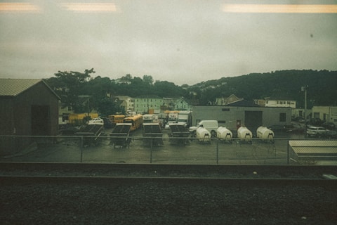 A fenced industrial area with several parked vehicles, including school buses, a FedEx truck, and white delivery vans. There are also utility trailers and equipment enclosed in tarp materials. Surrounding this are various buildings, including a warehouse, against a backdrop of green hills under an overcast sky.