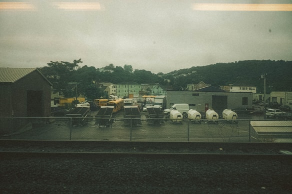 A fenced industrial area with several parked vehicles, including school buses, a FedEx truck, and white delivery vans. There are also utility trailers and equipment enclosed in tarp materials. Surrounding this are various buildings, including a warehouse, against a backdrop of green hills under an overcast sky.