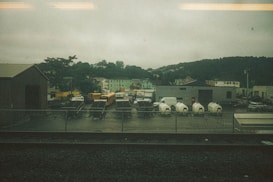 A fenced industrial area with several parked vehicles, including school buses, a FedEx truck, and white delivery vans. There are also utility trailers and equipment enclosed in tarp materials. Surrounding this are various buildings, including a warehouse, against a backdrop of green hills under an overcast sky.