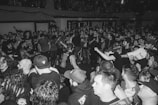 A large crowd of people gathered in a compact space, possibly a concert or music event. Most individuals are wearing dark clothing, with some displaying graphic t-shirts and band merchandise. The atmosphere appears lively and energetic, with people engaged in conversation and movement.