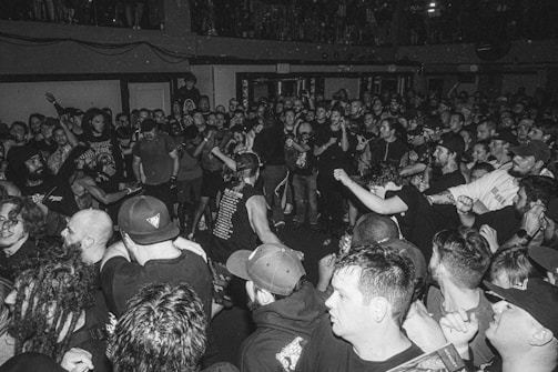 A large crowd of people gathered in a compact space, possibly a concert or music event. Most individuals are wearing dark clothing, with some displaying graphic t-shirts and band merchandise. The atmosphere appears lively and energetic, with people engaged in conversation and movement.