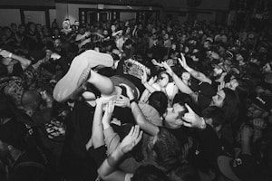 A black and white image showcases a large, energetic crowd at a music concert. People are tightly packed together with arms raised, and one person is crowd surfing. The scene is lively and dynamic, capturing the excitement and chaos typical of a live performance.