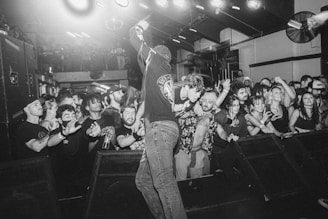 A lively concert scene with a singer engaging with an enthusiastic crowd. The audience is packed closely, showing expressions of enjoyment and excitement. Lighting fixtures and stage equipment are visible in the background, contributing to the vibrant atmosphere.