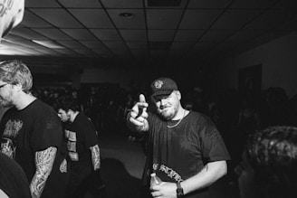 A black and white image features a man in a cap making a playful gesture towards the camera in a dimly lit room. The background includes other individuals, some of whom appear to have tattoos and are dressed casually. The environment gives the impression of a social gathering or event.