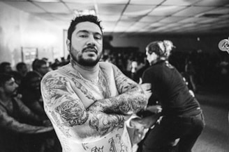 A heavily tattooed man with his arms crossed is prominently featured in the foreground. He has a serious expression and dark, short hair. In the background, there is a crowd of people, and another person who appears to be engaged in some energetic activity, possibly performing.