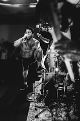 A dynamic black and white photograph captures a musician passionately performing on stage. The singer, heavily tattooed and shirtless, is energetically screaming into a microphone. One foot is propped up on a drum set, enhancing his intense stage presence. Other musicians and a drum kit are in the background, contributing to the lively atmosphere.