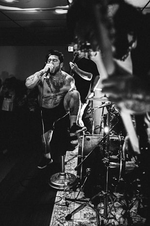 A dynamic black and white photograph captures a musician passionately performing on stage. The singer, heavily tattooed and shirtless, is energetically screaming into a microphone. One foot is propped up on a drum set, enhancing his intense stage presence. Other musicians and a drum kit are in the background, contributing to the lively atmosphere.