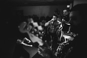 A high-energy live music performance with a vocalist passionately singing into a microphone. The scene is set in a dimly lit venue with an engaged audience in the background. Band members and musical instruments like a guitar and drums are visible, contributing to an intense and lively atmosphere.