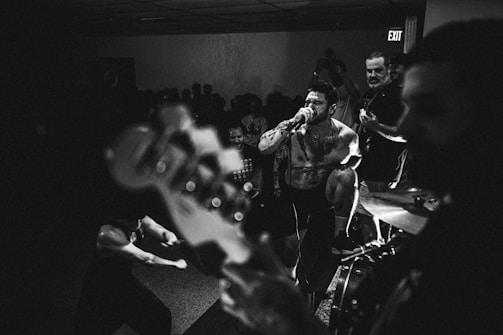 A high-energy live music performance with a vocalist passionately singing into a microphone. The scene is set in a dimly lit venue with an engaged audience in the background. Band members and musical instruments like a guitar and drums are visible, contributing to an intense and lively atmosphere.