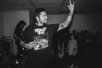 A black and white photo captures a tattooed man in a t-shirt with a graphic design, extending his arm and hand towards the audience. He stands in a room with a low ceiling, surrounded by a crowd of people. Another person appears in the background, suggesting a live performance setting.
