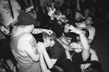 A black and white image of an energetic music concert featuring a crowd of enthusiastic fans and a musician performing on stage. The musician, with tattoos on their arms, holds a microphone and engages with the audience, who have their hands raised, showing excitement and engagement with the performance. The scene is packed with people, indicating a lively and boisterous atmosphere.
