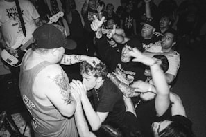 A black and white image of an energetic music concert featuring a crowd of enthusiastic fans and a musician performing on stage. The musician, with tattoos on their arms, holds a microphone and engages with the audience, who have their hands raised, showing excitement and engagement with the performance. The scene is packed with people, indicating a lively and boisterous atmosphere.