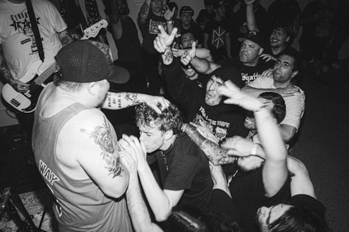 A black and white image of an energetic music concert featuring a crowd of enthusiastic fans and a musician performing on stage. The musician, with tattoos on their arms, holds a microphone and engages with the audience, who have their hands raised, showing excitement and engagement with the performance. The scene is packed with people, indicating a lively and boisterous atmosphere.