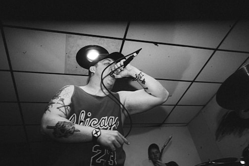 A black and white image of a man with tattoos performing in a small venue, holding a microphone close to his mouth. He is wearing a sleeveless jersey with the word 'Chicago' and the number '23'. Another musician is visible in the background, holding a guitar. The scene appears energetic, typical of a live performance.