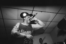 A black and white image of a man with tattoos performing in a small venue, holding a microphone close to his mouth. He is wearing a sleeveless jersey with the word 'Chicago' and the number '23'. Another musician is visible in the background, holding a guitar. The scene appears energetic, typical of a live performance.