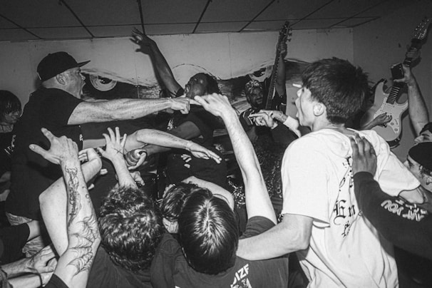 A black and white photograph captures a high-energy moment at a live music concert. Several people are in the crowd, reaching towards the stage where a musician is performing passionately. Two guitarists can be seen in the background. The people's expressions and actions convey excitement and engagement.