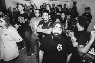 A group of people gathered closely in a room, many with tattoos and wearing casual clothing like t-shirts and caps. The photograph is black and white, giving it a vintage or nostalgic feel. The expressions range from neutral to mildly intense, suggesting they might be attending an event or concert.