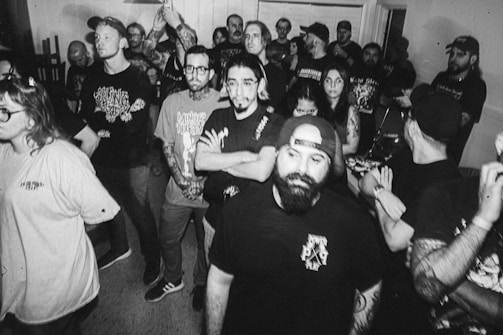 A group of people gathered closely in a room, many with tattoos and wearing casual clothing like t-shirts and caps. The photograph is black and white, giving it a vintage or nostalgic feel. The expressions range from neutral to mildly intense, suggesting they might be attending an event or concert.