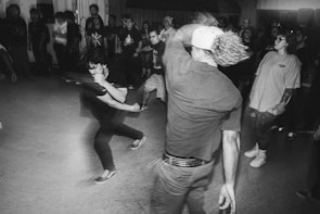 A lively indoor scene with a group of people energetically dancing and moving around in a crowded room, lit with dim lighting. The movement is dynamic, with people wearing casual clothing and some observers watching from the side.