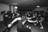 A group of young people are energetically dancing in a room with a crowd watching. The atmosphere is lively with individuals expressing themselves through vigorous dance movements, typical of a rock or punk concert.