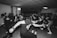 A group of young people are energetically dancing in a room with a crowd watching. The atmosphere is lively with individuals expressing themselves through vigorous dance movements, typical of a rock or punk concert.
