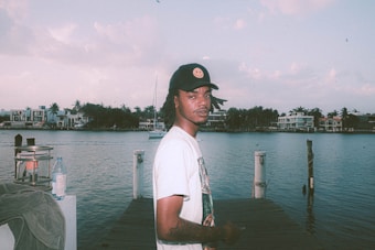 A person stands on a wooden dock in front of a calm body of water, with boats and large houses in the background. They wear a black cap with a smiley face and a printed t-shirt. There's a lamp and a bottle on the dock.
