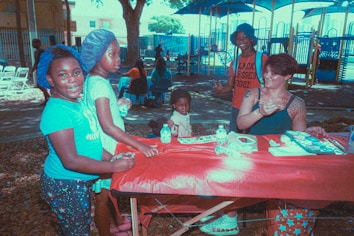 A group of people gather around a table covered with a red cloth in an outdoor setting, possibly a park. Children and an adult are present, engaging in a creative activity involving painting. A woman with tattoos on her arm assists the children with the task. In the background, there are playground structures and other people seated.